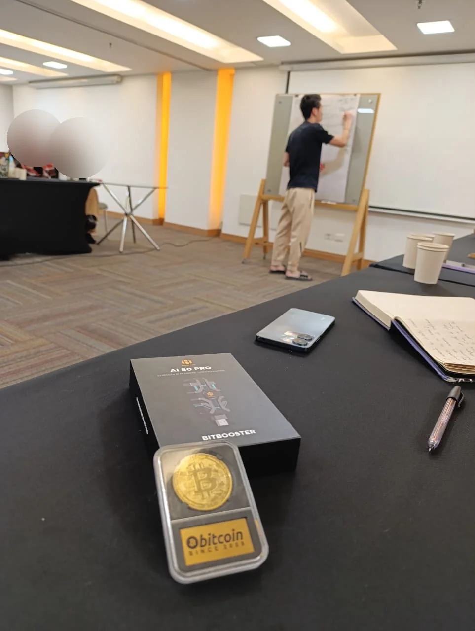 Hugo Suah de espaldas presentando BitHarvest en una reunión, con una caja del dispositivo BitBooster sobre la mesa